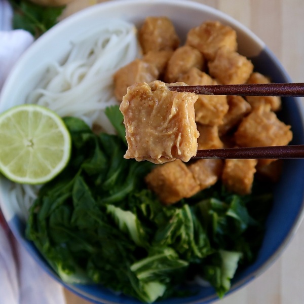 Bowl with rice noodles, bok choy, and peanut tofu with chopsticks holding up a piece of peanut tofu.