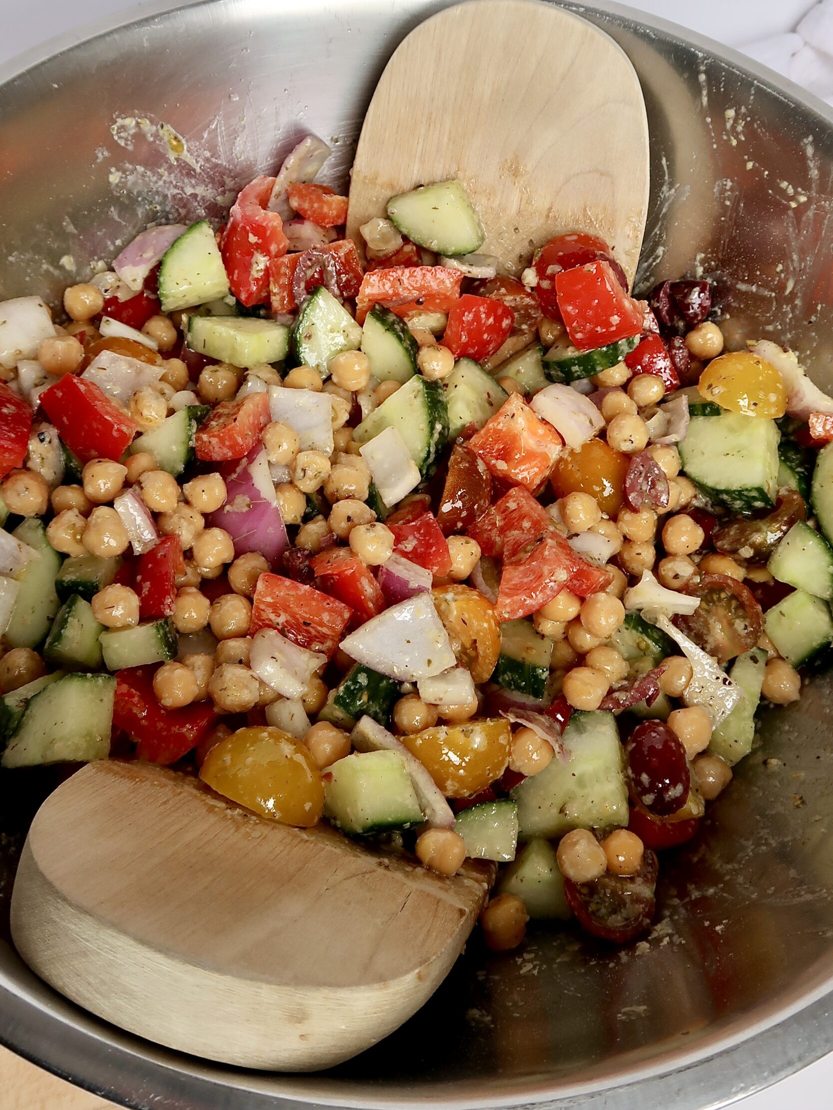Metal bowl with cucumber, kalamata olives, cherry tomatoes, red onions and chickpeas inside tossed in dressing.