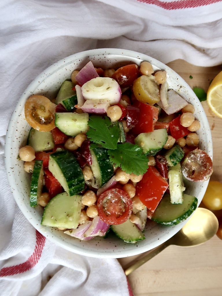 White bowl with cherry tomatoes, cucumbers, chickpeas, kalamata olives and dressing in it.