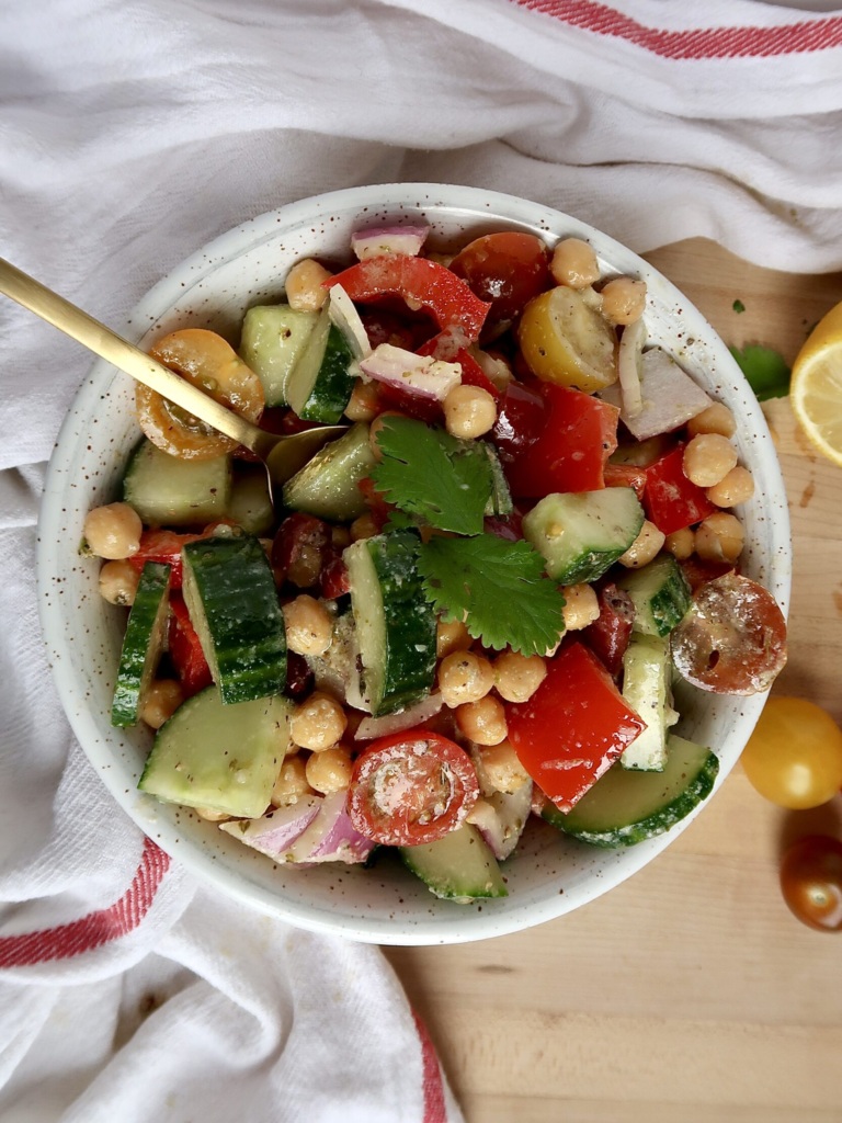 White bowl with chickpeas, tomatoes, cucumber, bell pepper inside and a spoon with a jar labeled 'Greek salad dressing' and a lemon on the side.