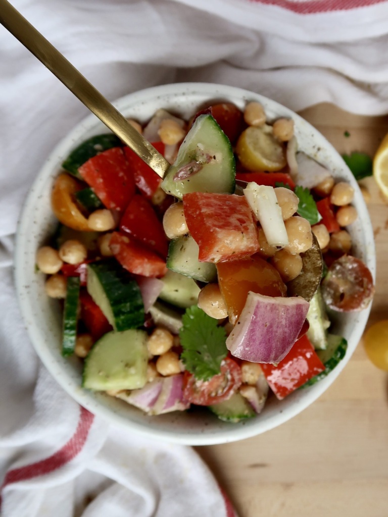 White bowl with chickpeas, tomatoes, cucumber, bell pepper, with a spoon inside.