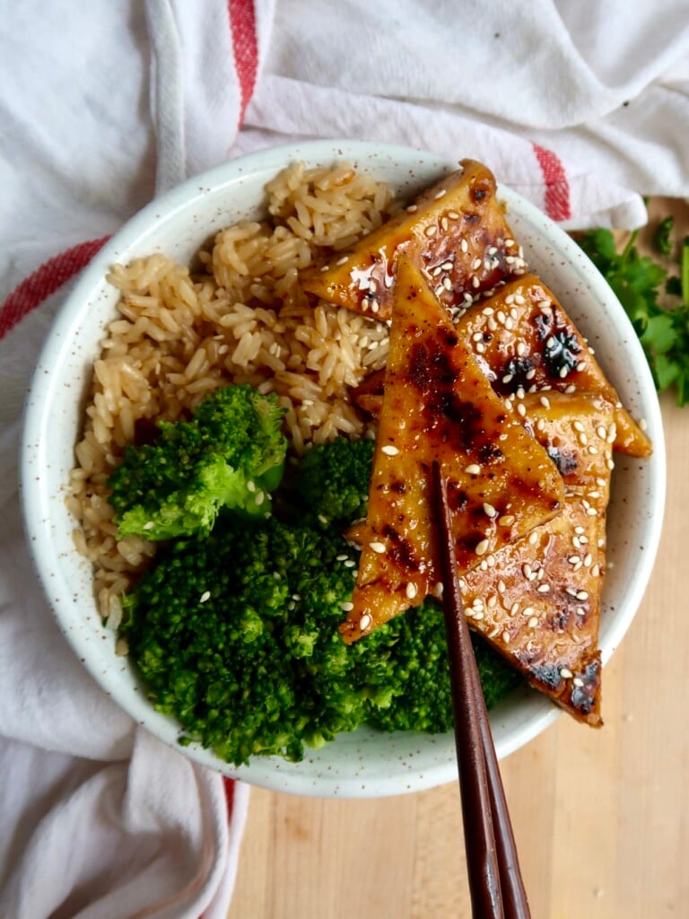 Brown rice, broccoli and maple glazed tofu in a bowl with sesame seeds on top with a pair of chopsticks holding them up.