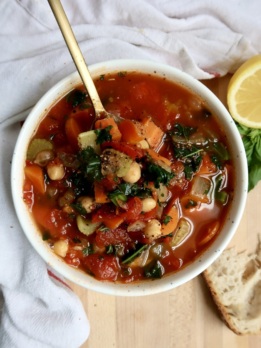Bowl with veggie soup in it with a spoon as well and bread on the side.