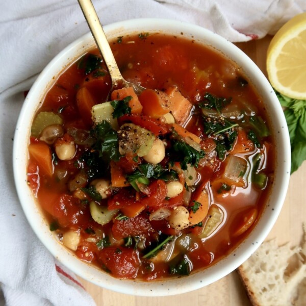 Bowl with veggie soup in it with a spoon as well and bread on the side.