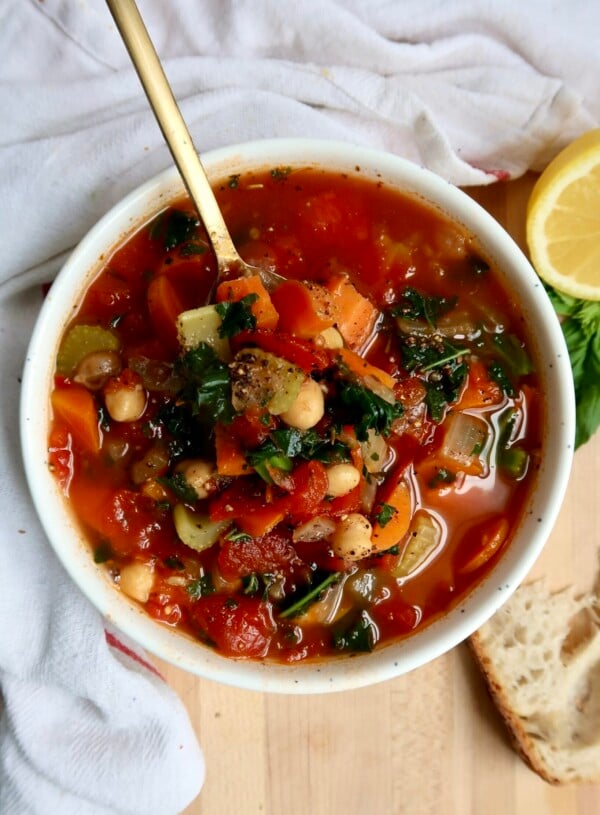 Bowl with veggie soup in it with a spoon as well and bread on the side.