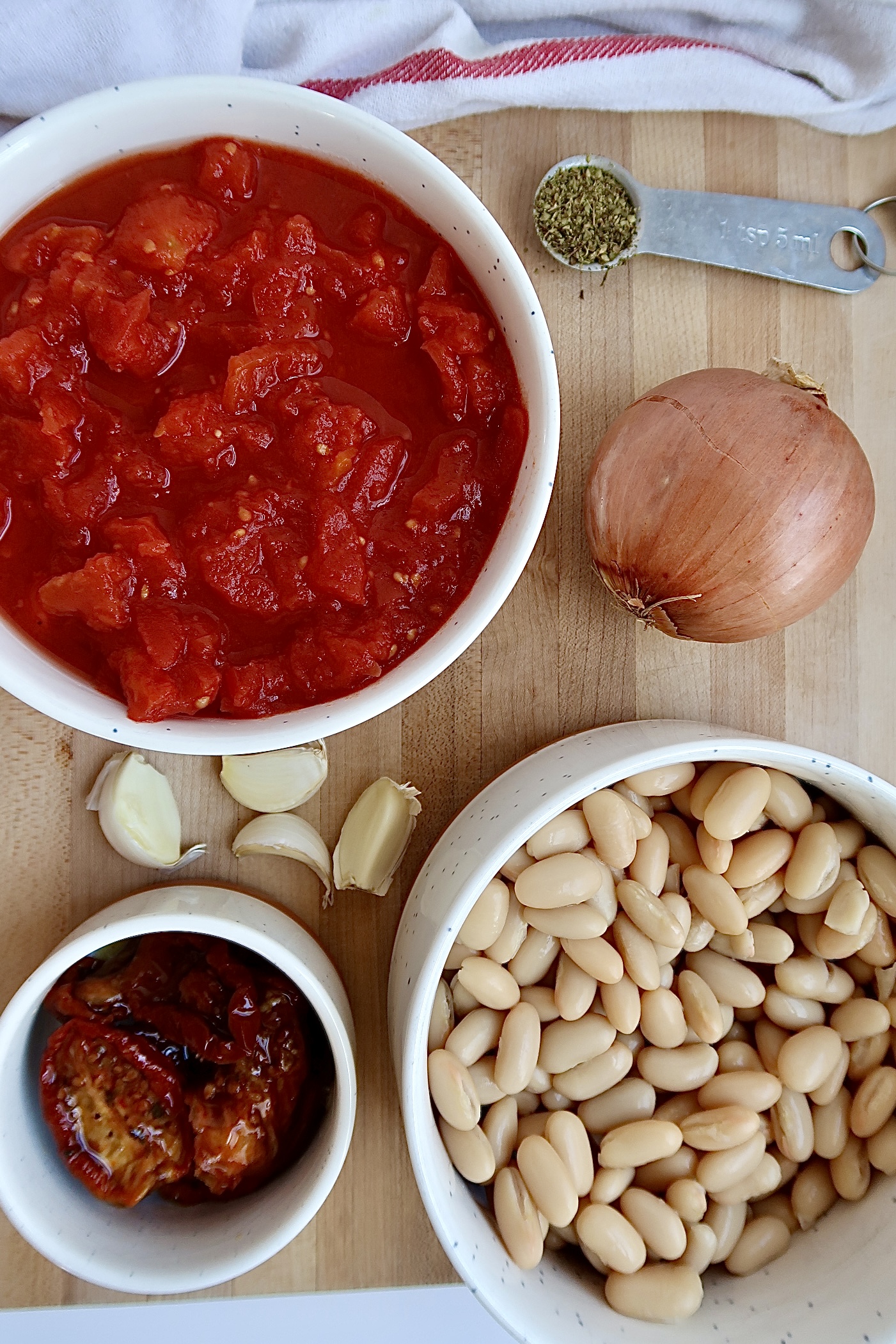 Cutting board with a white bowl with diced tomatoes in it, with an onion, garlic, a hwite bowl with white beans and sundried tomatoes on the side.