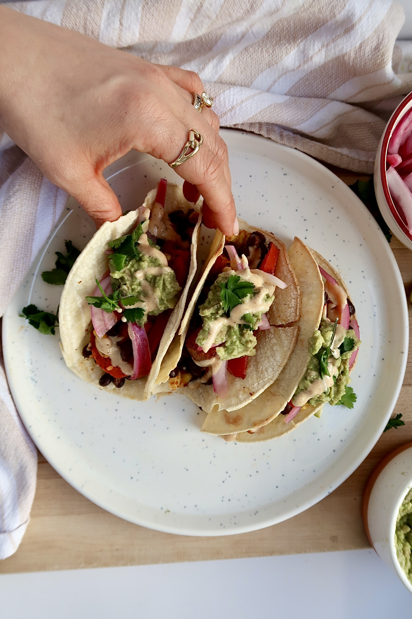 Three tacos with black beans, red onion, avocado, bell pepper and cilantro with a hand grabbing one.