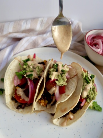Three tacos with black beans, red onion, avocado, bell pepper and cilantro with a chipotle sauce being drizzled on top.