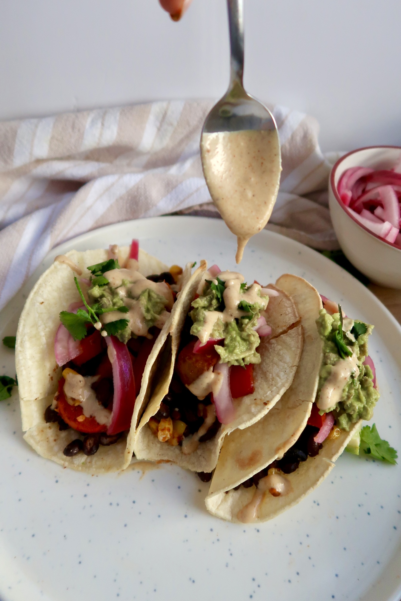 Three tacos with black beans, red onion, avocado, bell pepper and cilantro with a chipotle sauce being drizzled on top.