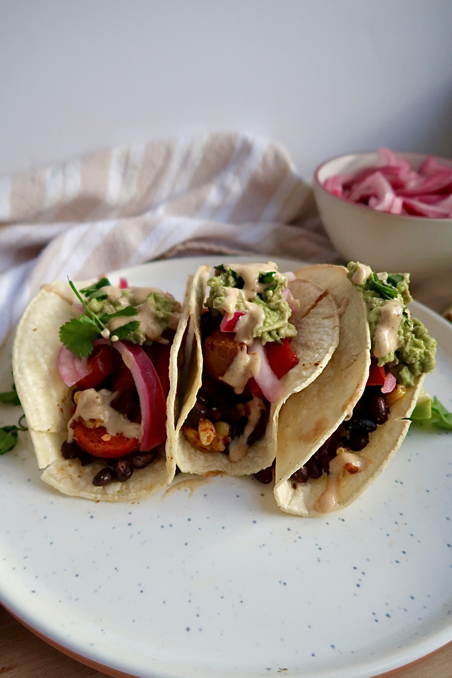Side view of Three tacos with black beans, red onion, avocado, bell pepper and cilantro.