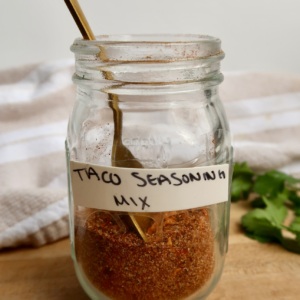 Small clear jar labeled 'taco seasoning mix' with spices in it and a spoon.