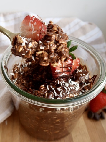 Brownie batter overnight oats in a mason jar with strawberries and coconut flakes on top and a spoon scooping some.