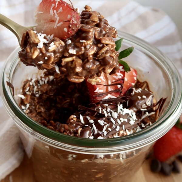 Brownie batter overnight oats in a mason jar with strawberries and coconut flakes on top and a spoon scooping some.