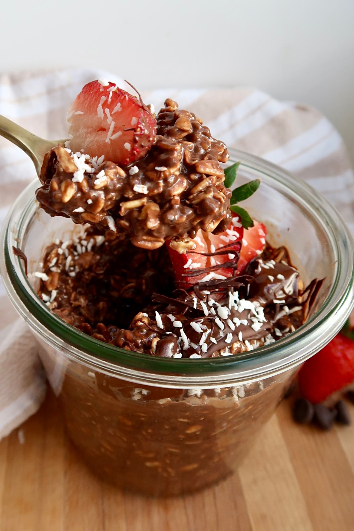 Brownie batter overnight oats in a mason jar with strawberries and coconut flakes on top and a spoon scooping some.