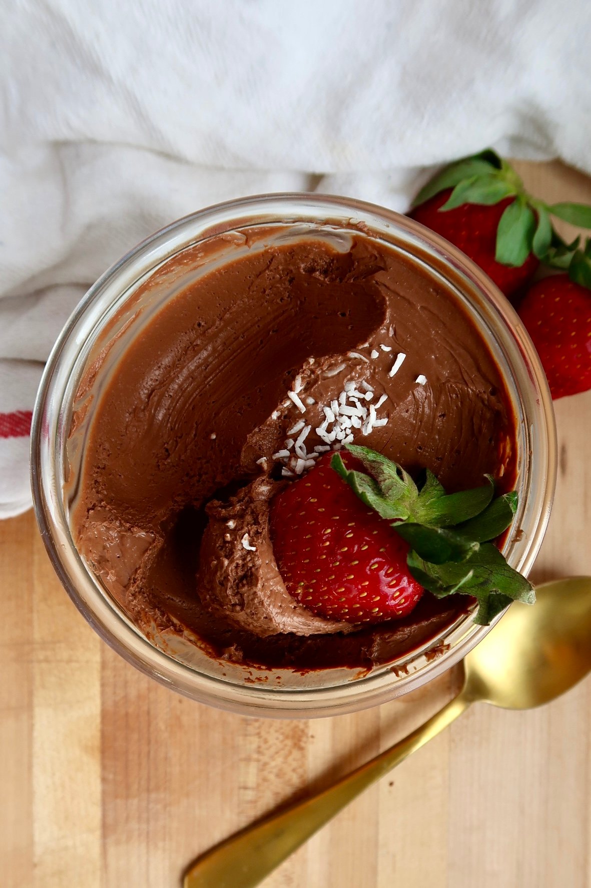 Overhead shot of Jar of chocolate mousse with coconut flakes and a strawberry on top with some of the mousse scooped out and strawberry being dipped.