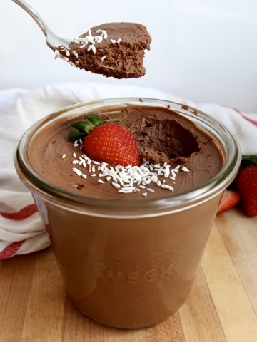 Side view of Jar of chocolate mousse with coconut flakes and a strawberry on top with a spoon scooping some of the mousse.