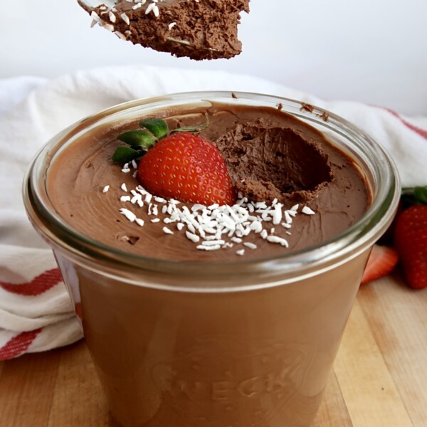 Side view of Jar of chocolate mousse with coconut flakes and a strawberry on top with a spoon scooping some of the mousse.