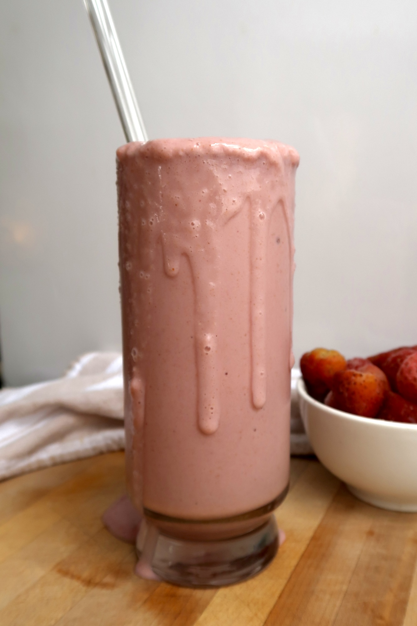 Glass with a strawberry smoothie dripping off the side with a bowl of strawberries to the side.