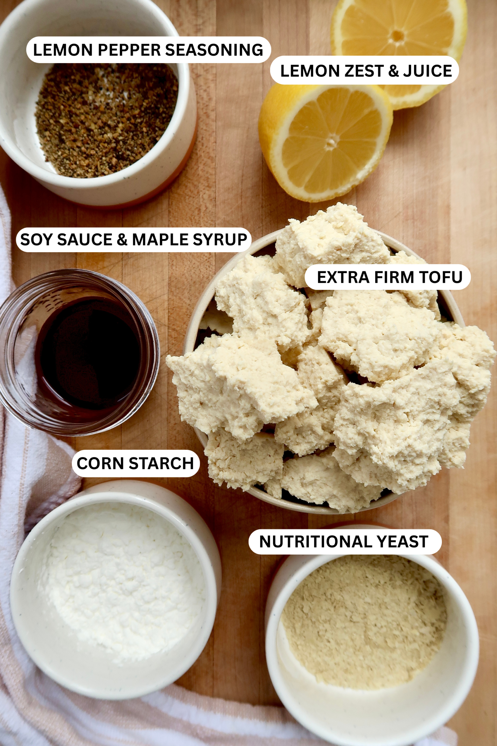 Cutting board with torn tofu, lemon, lemon pepper seasoning, nutritional yeast, soy sauce and corn starch on it, labeled.