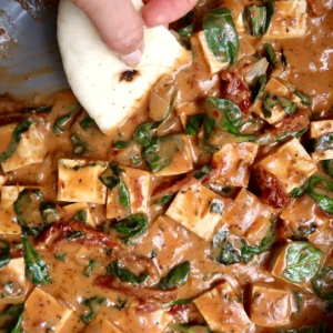 Cubed tofu in a pan with a sauce and spinach wilted in and naan bread scooping some up.
