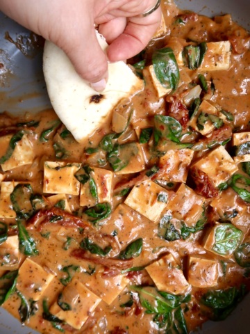 Cubed tofu in a pan with a sauce and spinach wilted in and naan bread scooping some up.