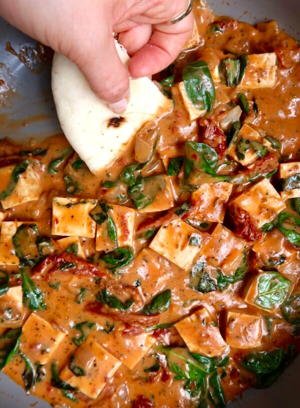 Cubed tofu in a pan with a sauce and spinach wilted in and naan bread scooping some up.