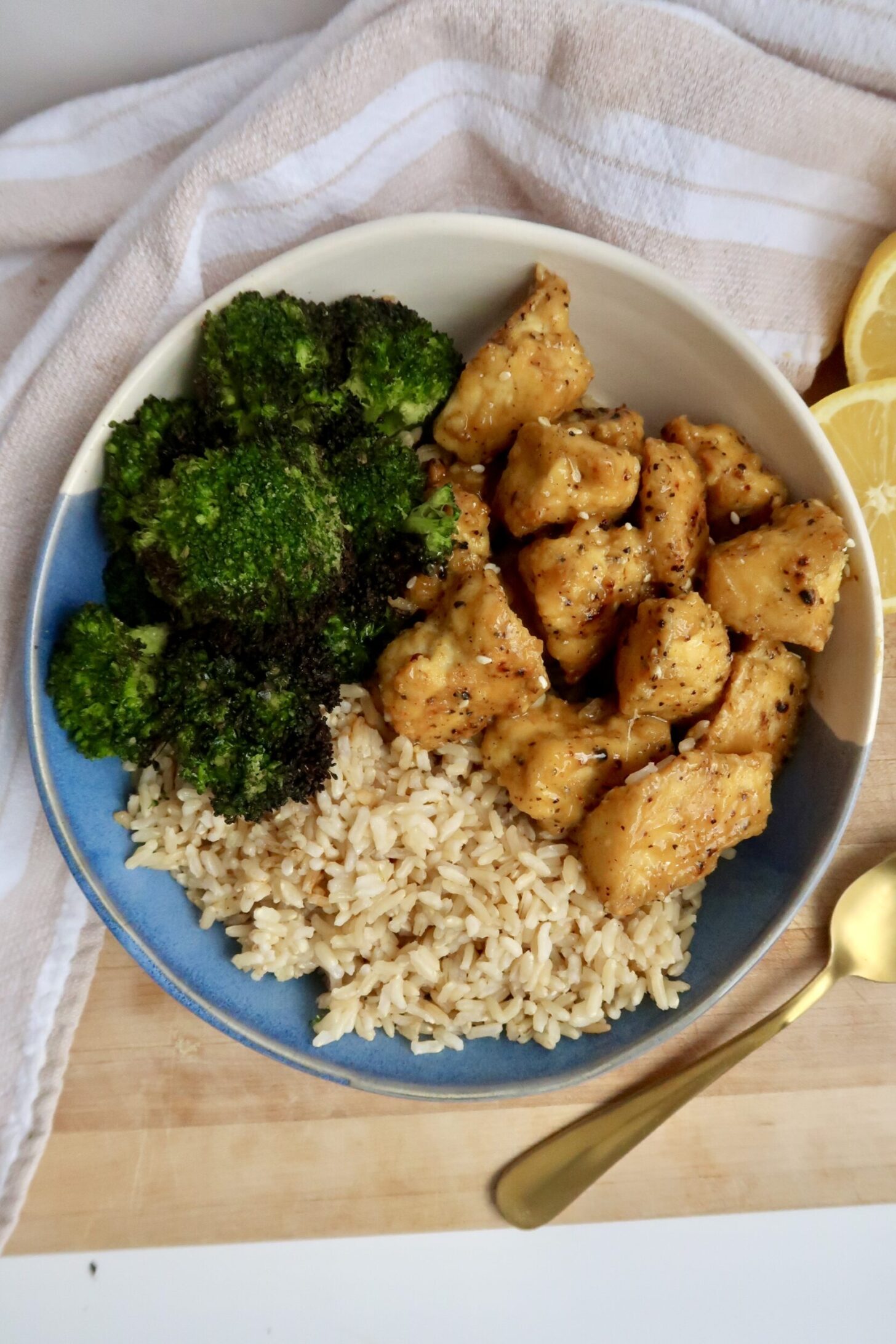 Tofu with a crispy coating and tossed in lemon pepper seasoning in a bowl with broccoli, brown rice and lemon to the side.