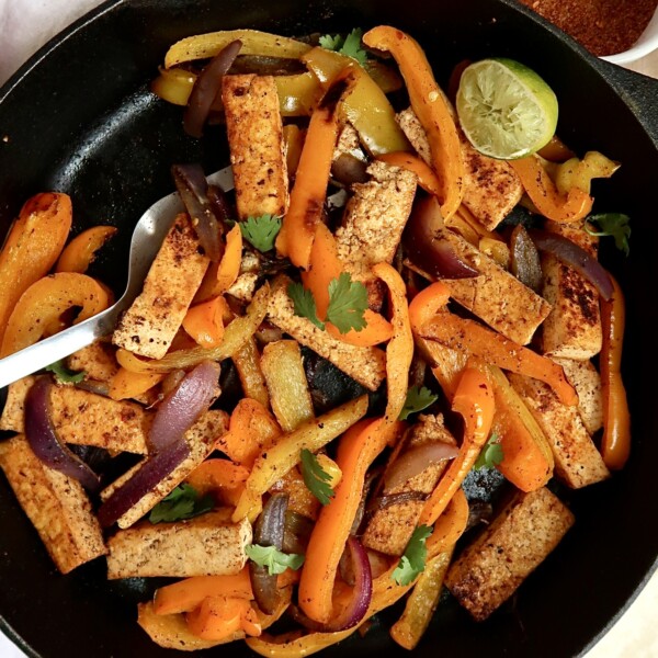 Pan of tofu fajita filling with peppers and a spatula and seasoning mix to the side.
