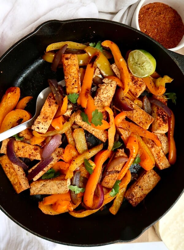 Pan of tofu fajita filling with peppers and a spatula and seasoning mix to the side.