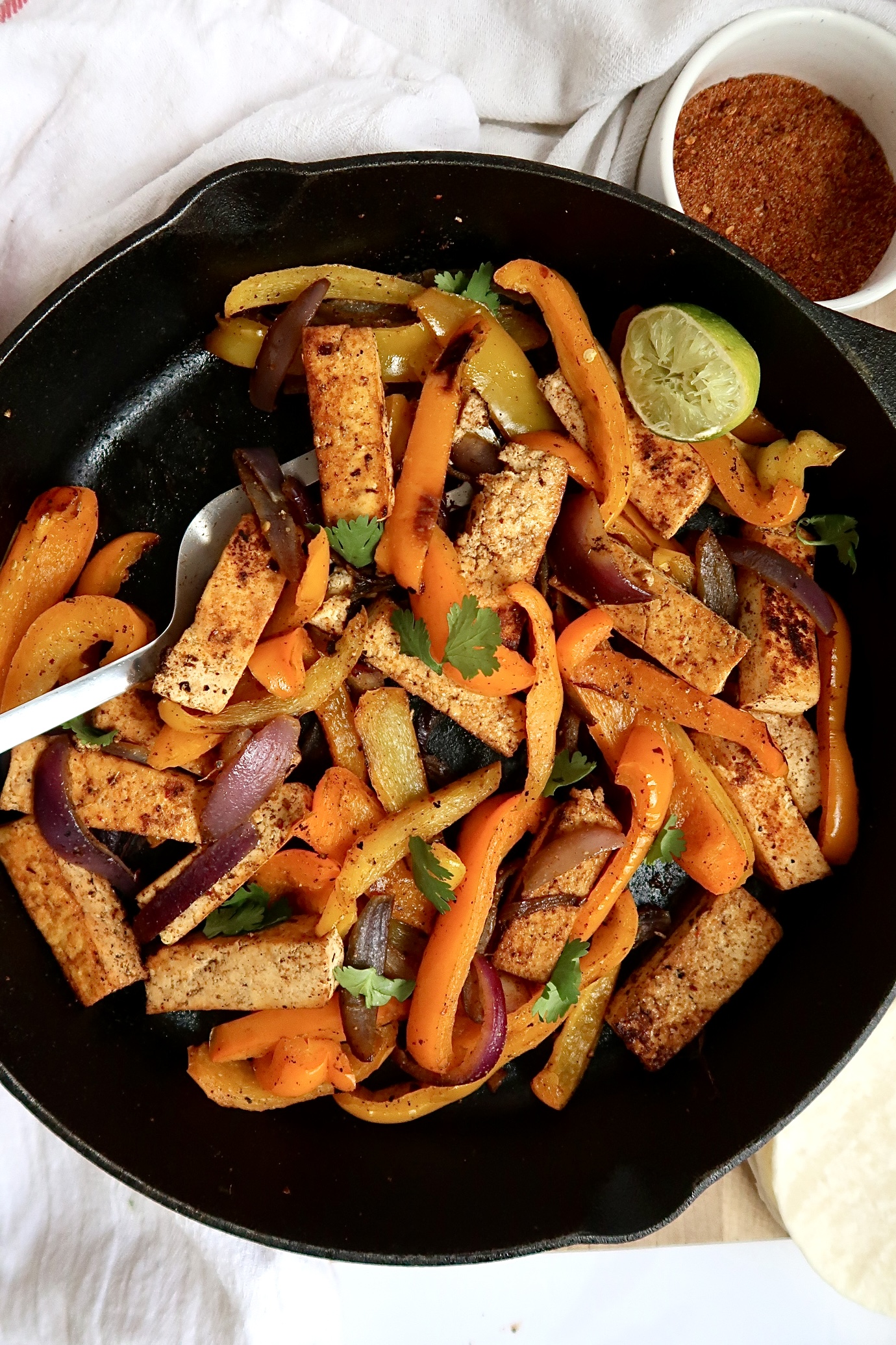 Pan of tofu fajita filling with peppers and a spatula and seasoning mix to the side. 
