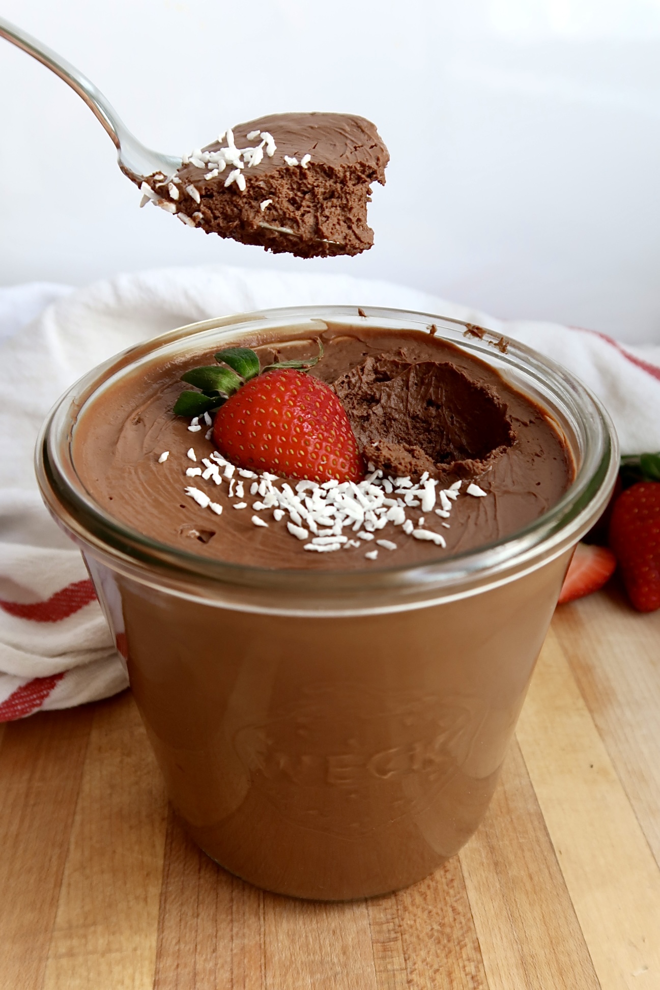 Side view of Jar of chocolate mousse with coconut flakes and a strawberry on top with a spoon scooping some of the mousse. 