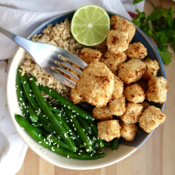 Bowl of coconut tofu with green beans, rice and lime to the side and a fork with a piece of tofu.