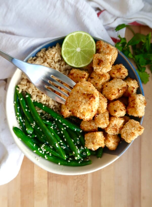 Bowl of coconut tofu with green beans, rice and lime to the side and a fork with a piece of tofu.