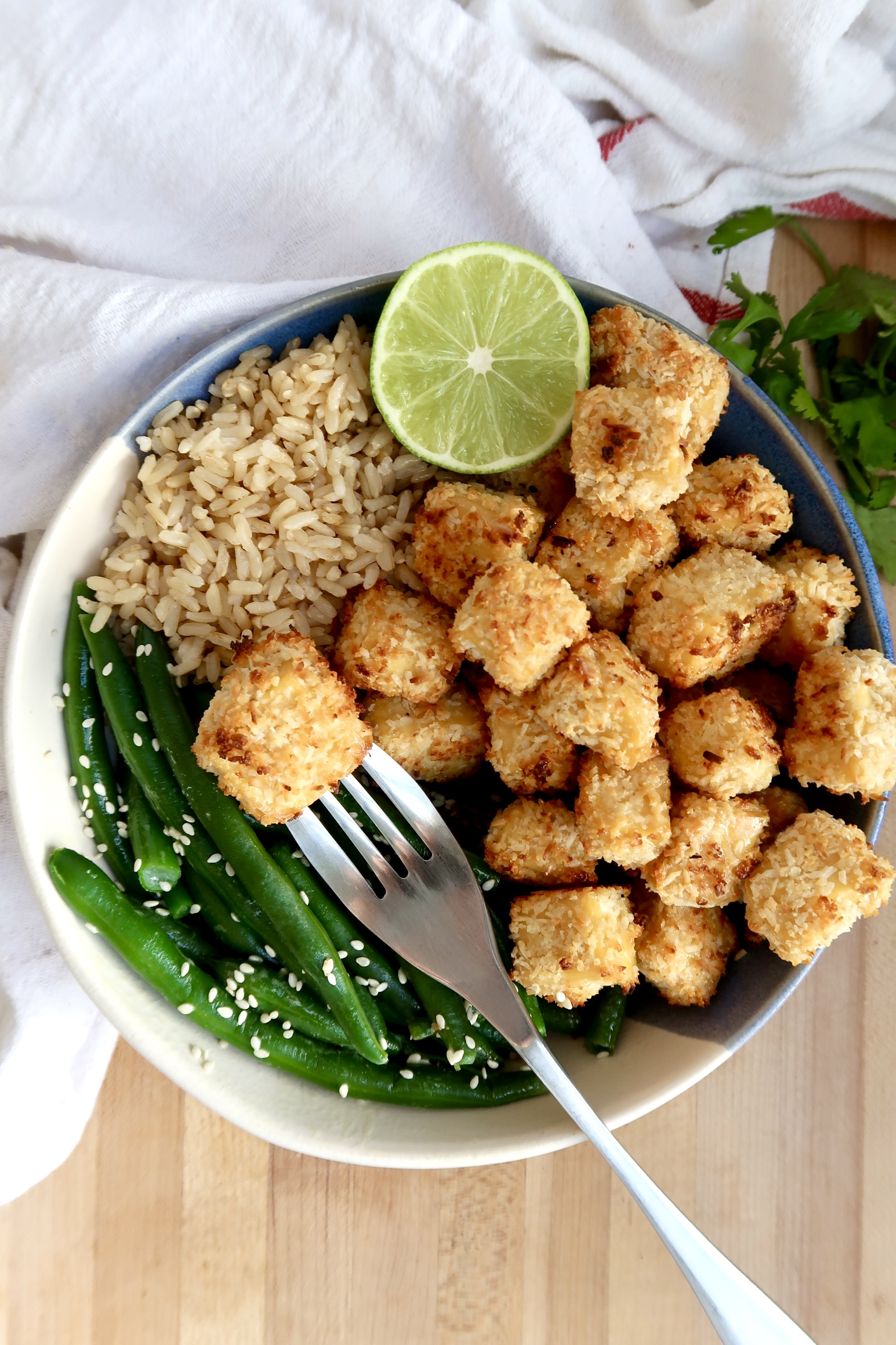 Bowl of coconut tofu with green beans, rice and lime to the side and a fork with a piece of tofu.