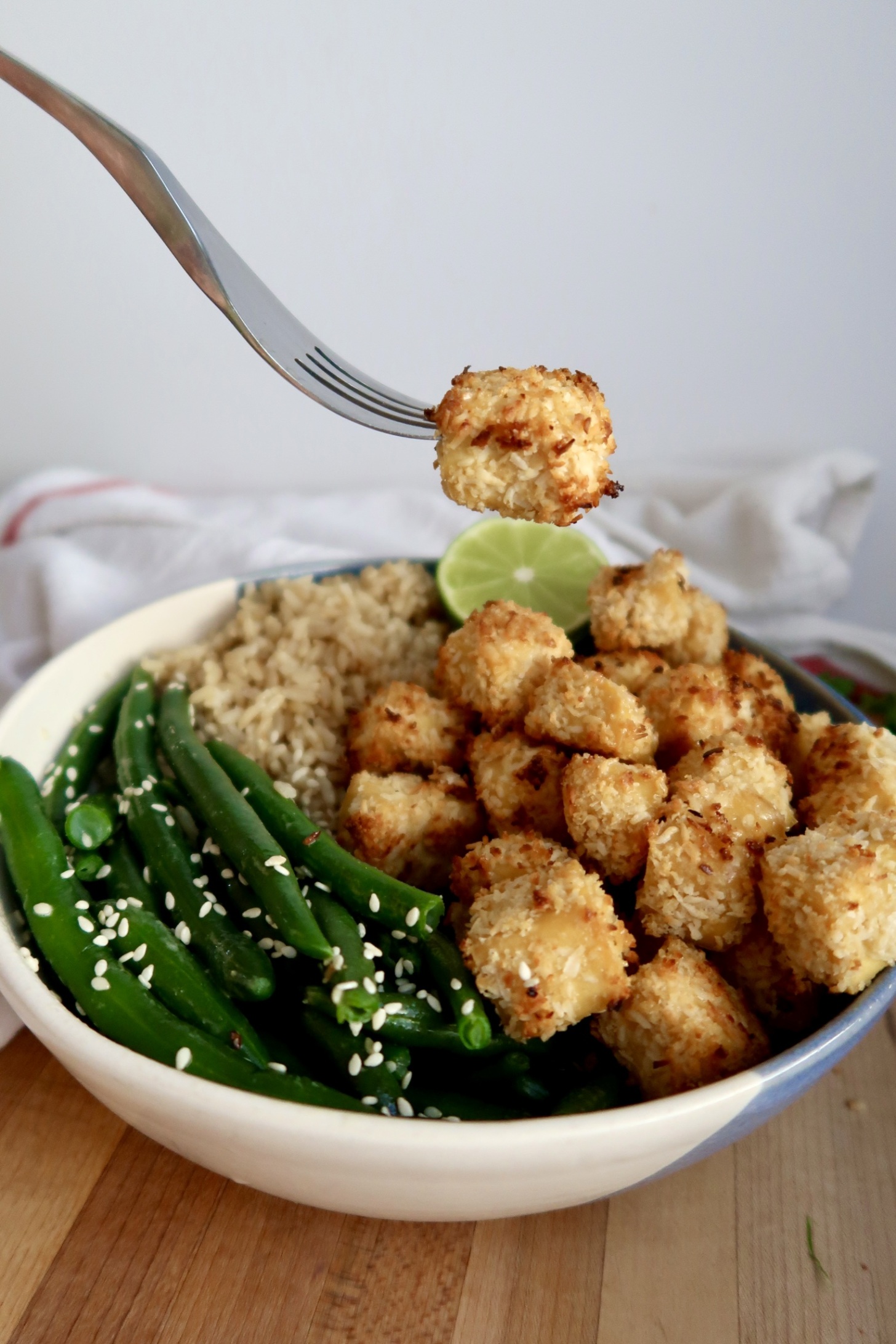 Side view of bowl of coconut tofu with green beans, rice and lime to the side and a fork with a piece of tofu in the air.