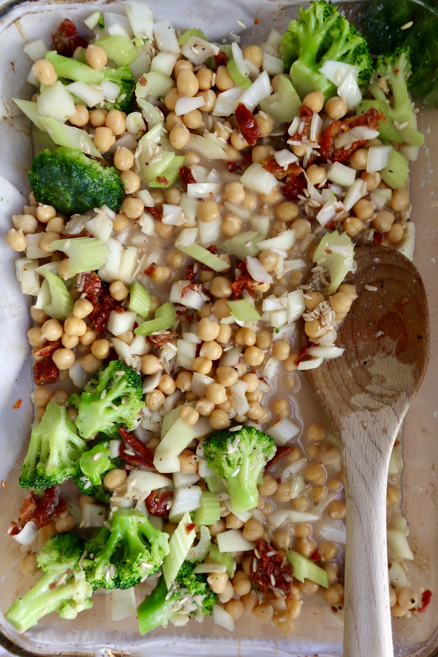 Casserole dish with rice, chickpeas, broccoli, celery and sundried tomatoes mixed in with a spoon.