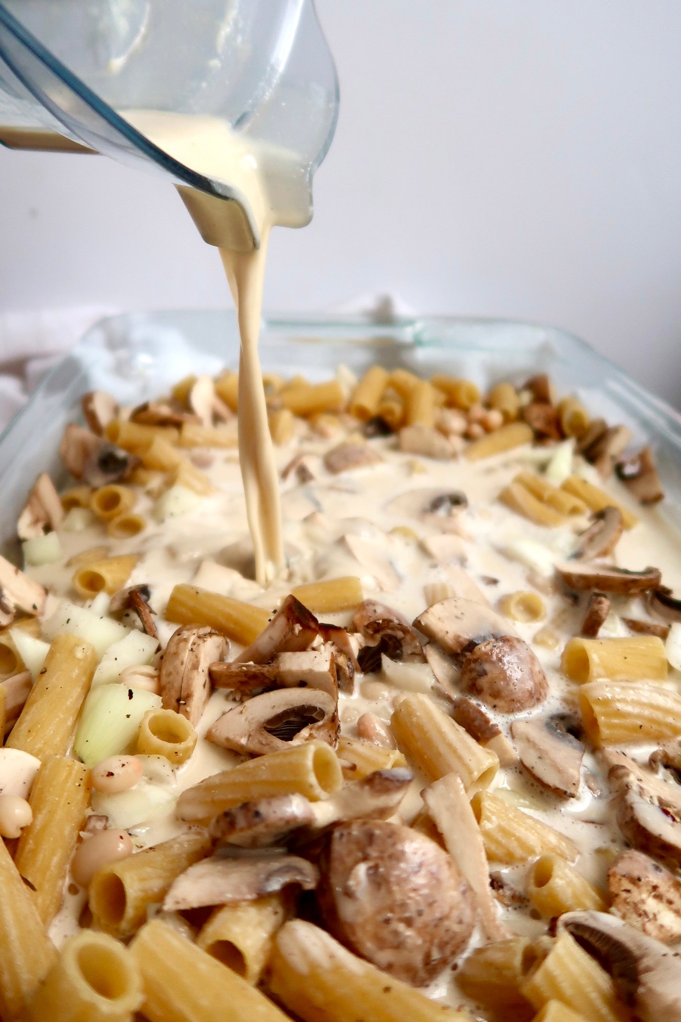 Casserole dish with pasta, mushrooms, diced onion and a creamy sauce being poured in.