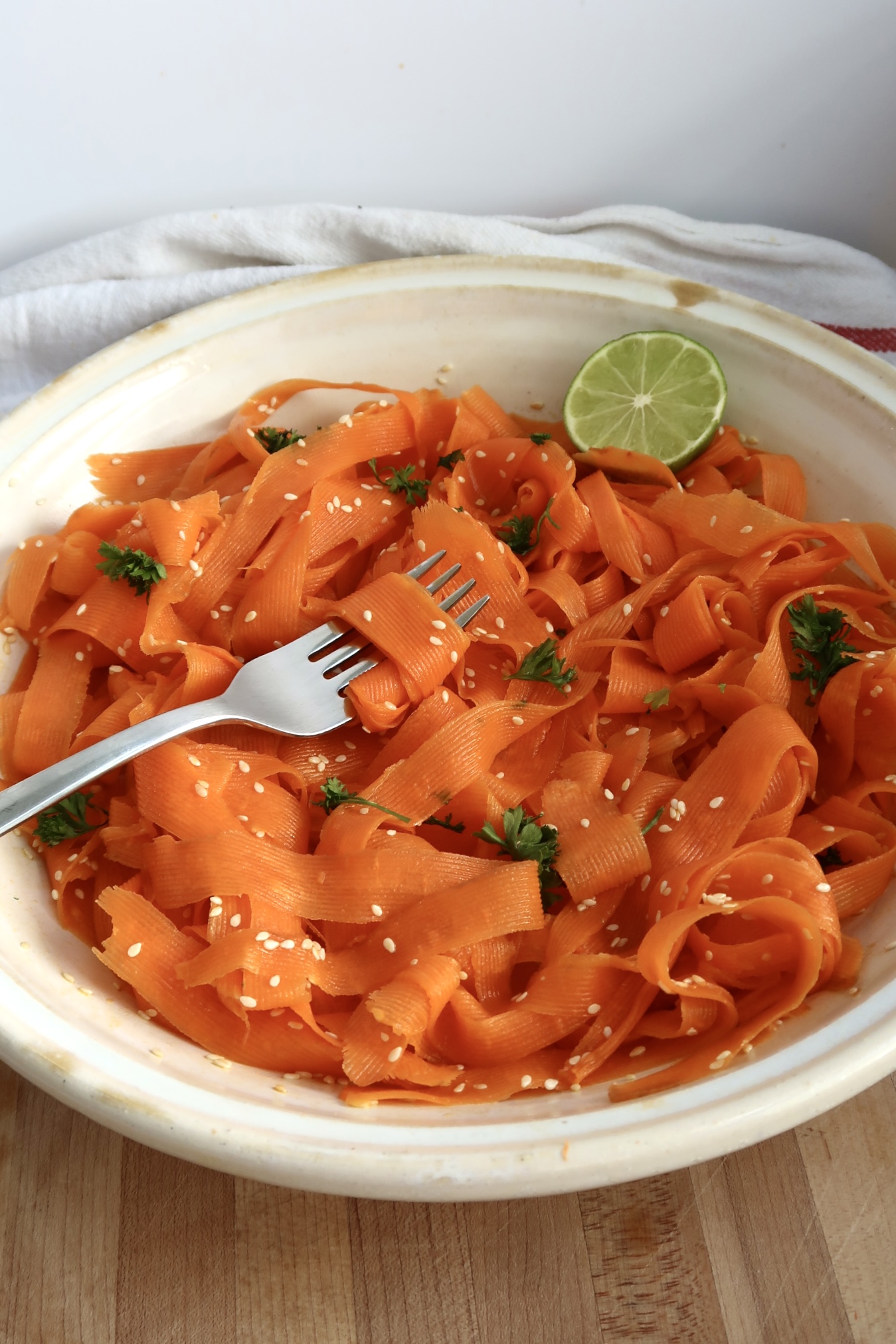 Side view of shaved carrots with sesame seeds, parsley and lime with a fork spiraling some.