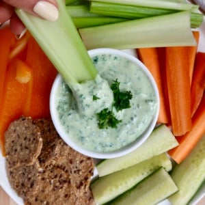 Hand holding celery being dipped in tofu dip with other veggies around.