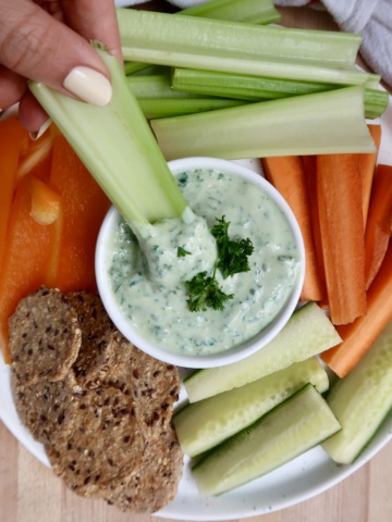 Hand holding celery being dipped in tofu dip with other veggies around.