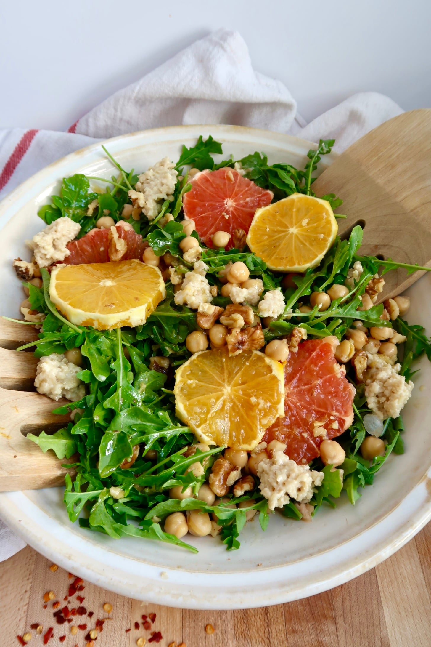 Arugula salad with citrus on top and ricotta and chickpeas and salad dressing to the side and salad tongs inside.
