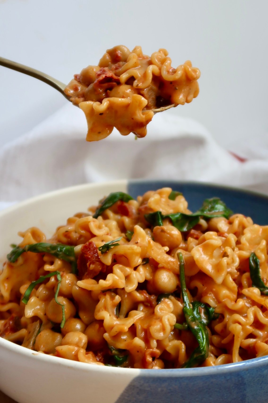Bowl with pasta in it with spinach, sundried tomatoes and chickpeas with a spoon scooping some.