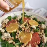 Arugula salad with citrus on top and ricotta and chickpeas and salad dressing being poured on top.