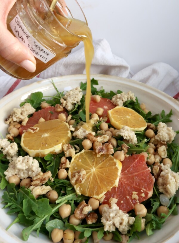 Arugula salad with citrus on top and ricotta and chickpeas and salad dressing being poured on top.
