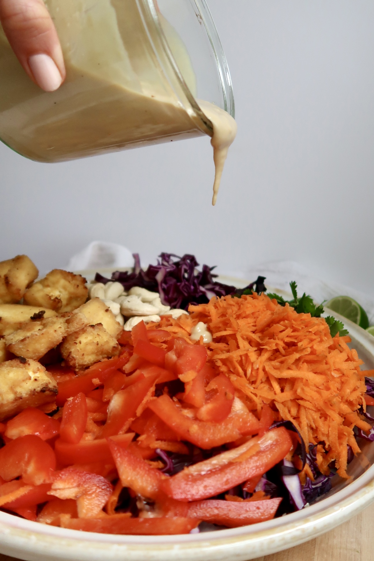 Dressing being poured over a bowl of tofu, shredded carrots, red pepper, cabbage, and cashews.