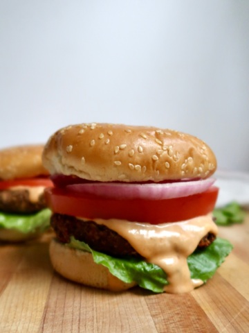 Black bean burger topped with lettuce, tomato, onion, and a generous drizzle of sauce on a sesame bun.
