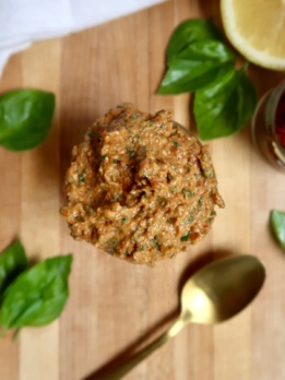 Large spoonful of sundried tomato pesto held over a wooden surface with fresh basil and a gold spoon nearby.
