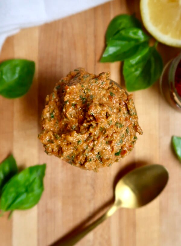Large spoonful of sundried tomato pesto held over a wooden surface with fresh basil and a gold spoon nearby.