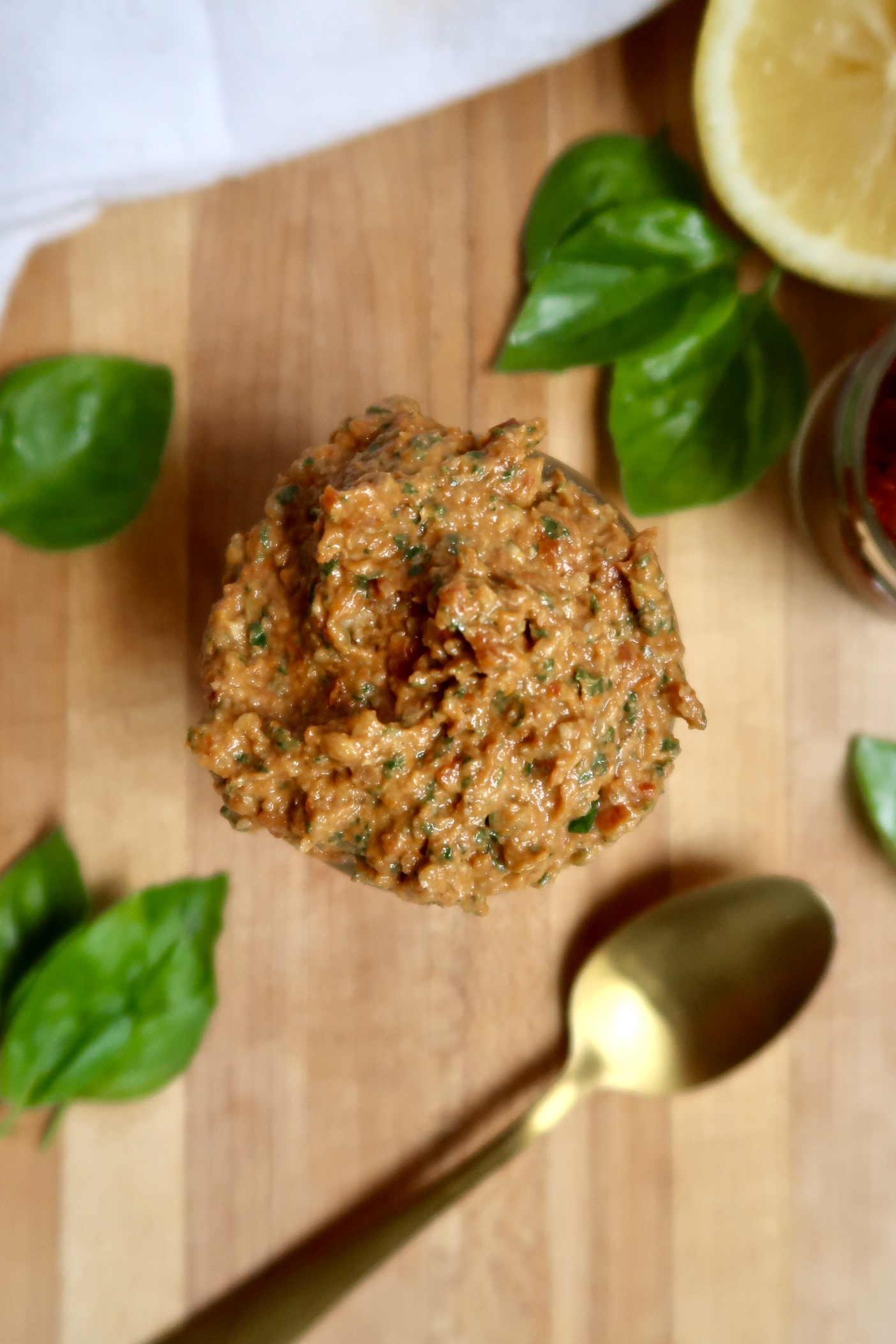 Large spoonful of sundried tomato pesto held over a wooden surface with fresh basil and a gold spoon nearby.