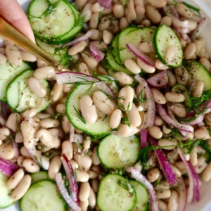 Spoon scooping a portion of the bean salad from the bowl, showcasing white beans, cucumber slices, red onion, and dill.